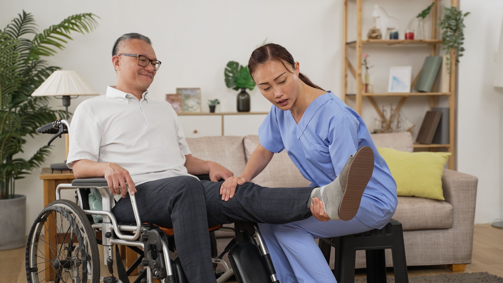 asian female care attendant helping senior man on wheelchair doing rehab training. she slowly lifts his injured leg while he shows painful face in the living room at home